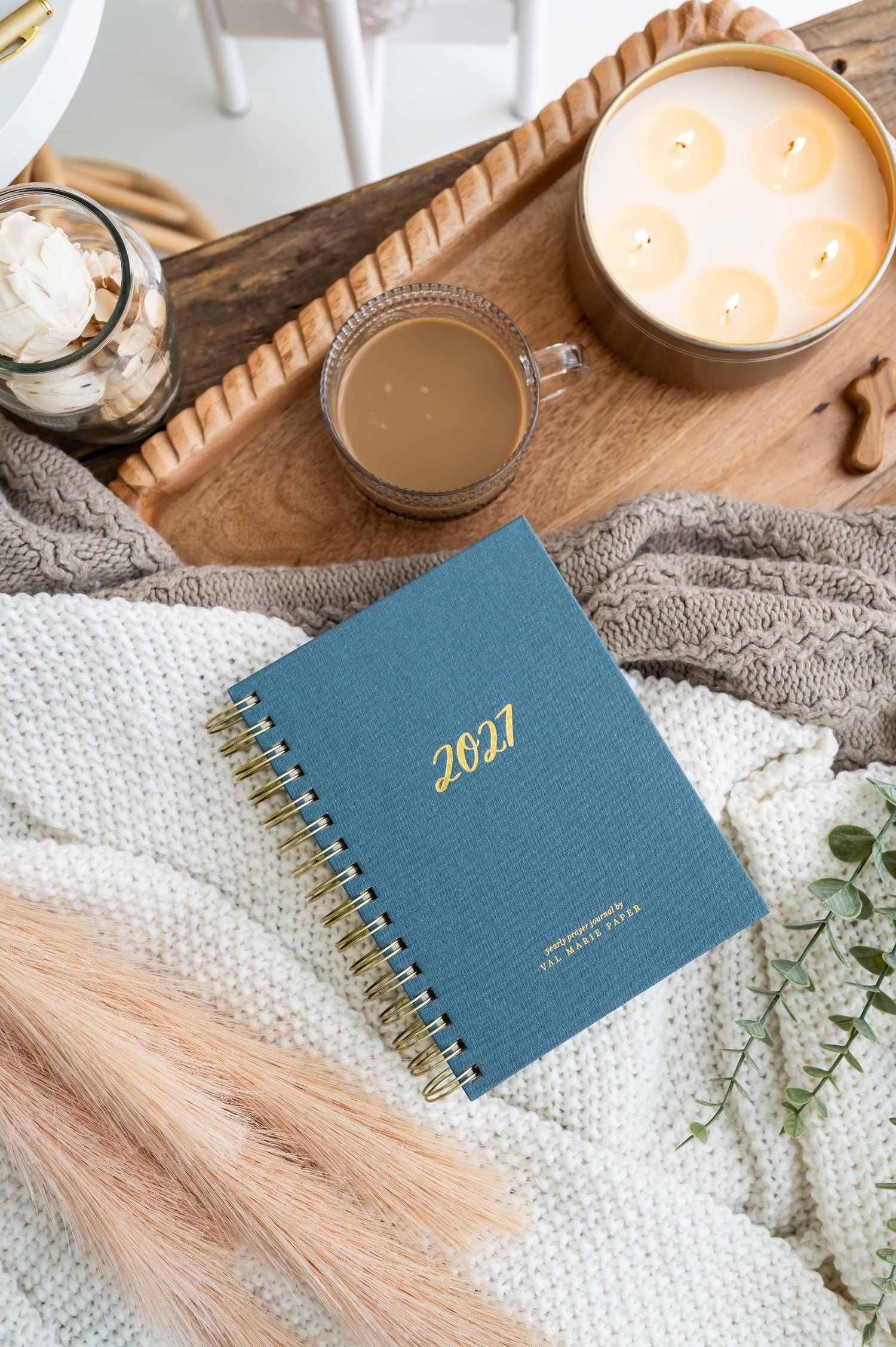 A blue 2027 spiral-bound prayer journal lies on a white knit blanket on a wooden tray, styled with a glass mug of coffee, a lit candle, and dried grasses.